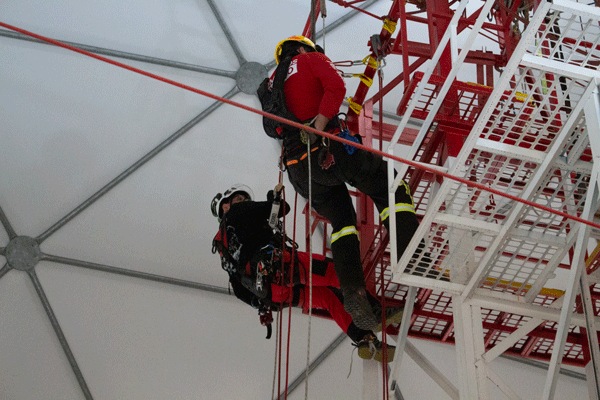 Dos alumnos con arnés y casco suspendidos de cuerdas junto a una estructura roja de entrenamiento dentro del domo de capacitación de Macaya Safety